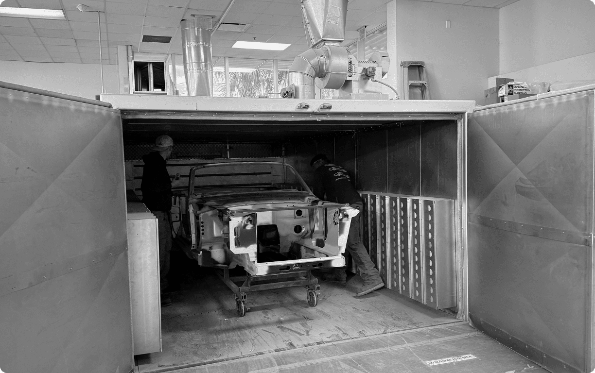 Black and white photograph of a classic Ford Mustang body shell inside an industrial curing oven for heat-setting structural adhesives at a professional restomod facility.