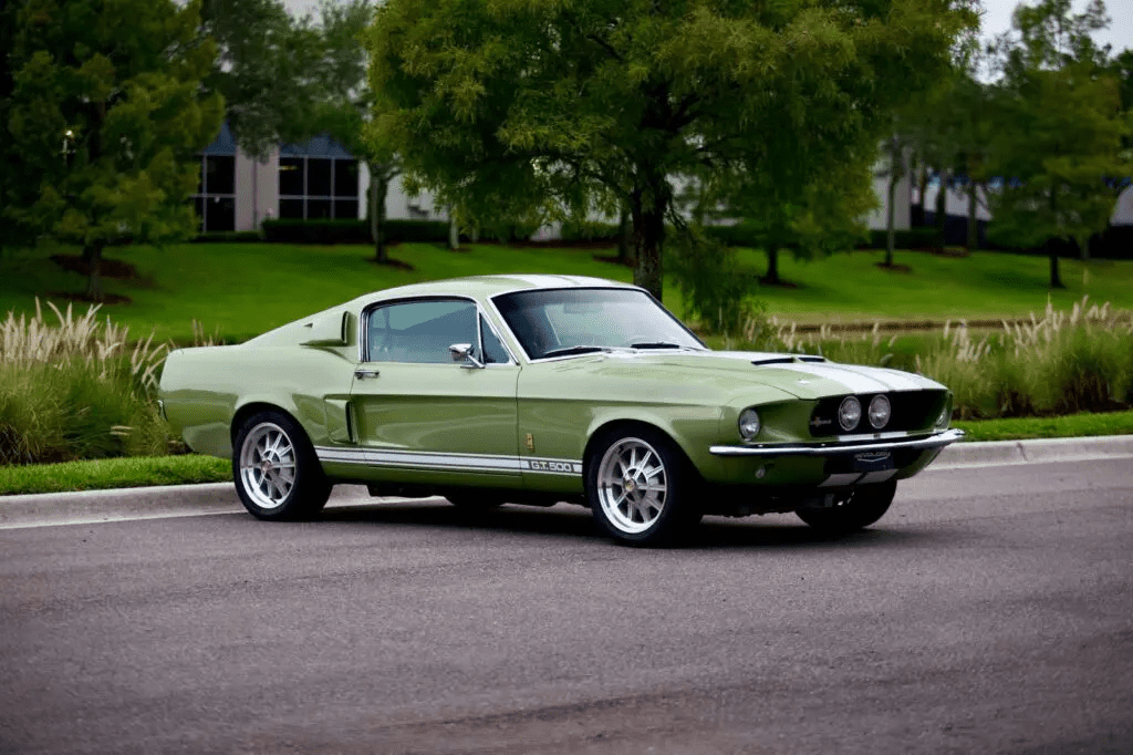 Green 1967 Revology Shelby GT500 Fastback parked on a tree-lined road in a front three-quarter view.