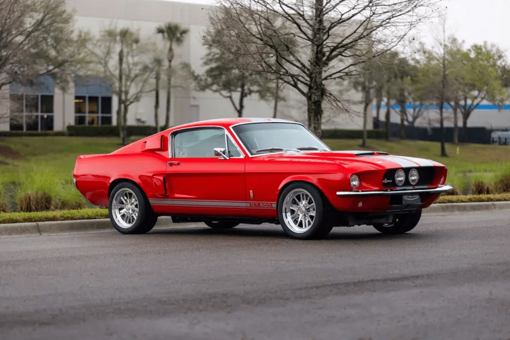 Red 1967 Revology Shelby GT500 Fastback with white racing stripes parked on a road in a front three-quarter view.