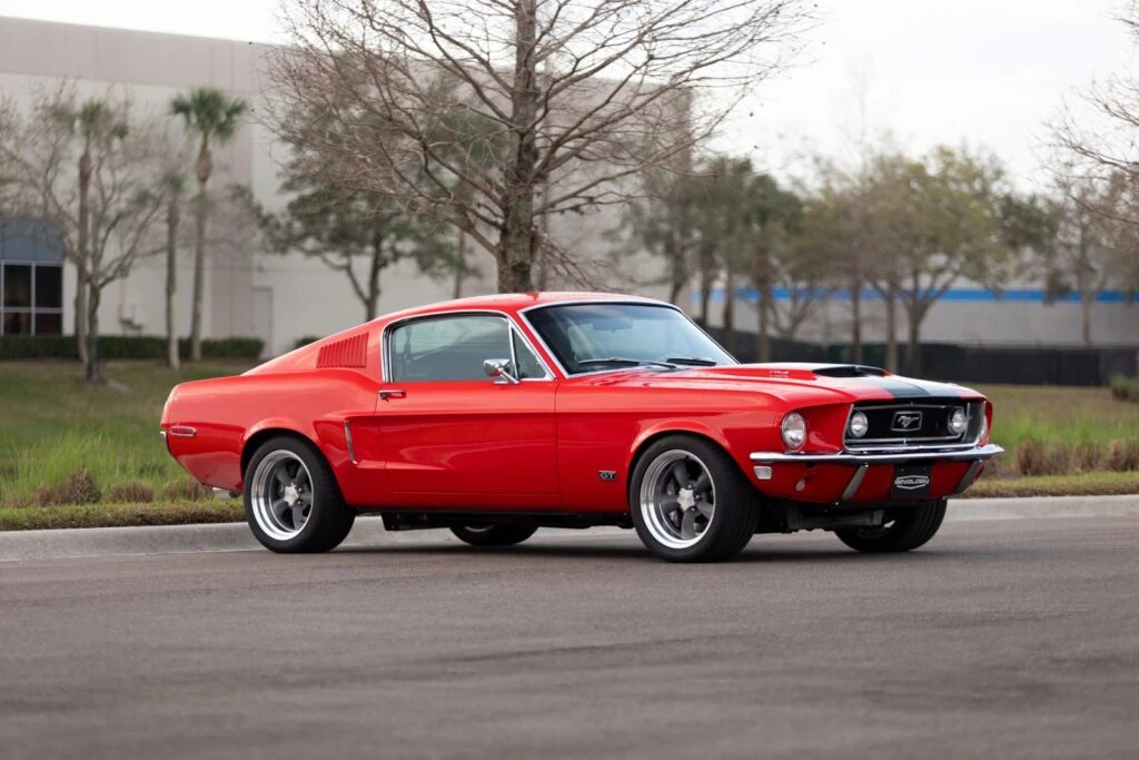 Red 1968 Revology Mustang GT Fastback Cobra Jet with a black hood scoop, parked on a road in a front three-quarter view.