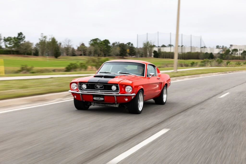 Red 1968 Revology Mustang GT Fastback Cobra Jet with a black hood scoop driving on an open road in a front three-quarter view.
