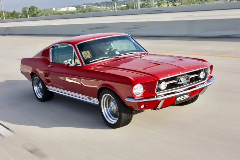 Red 1967 Ford Mustang fastback driving on a highway with classic chrome trim